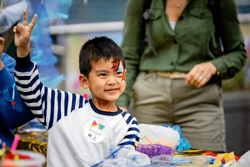 Young child wearing face paint stands making a peace sign with their hands during a crafting activity. 