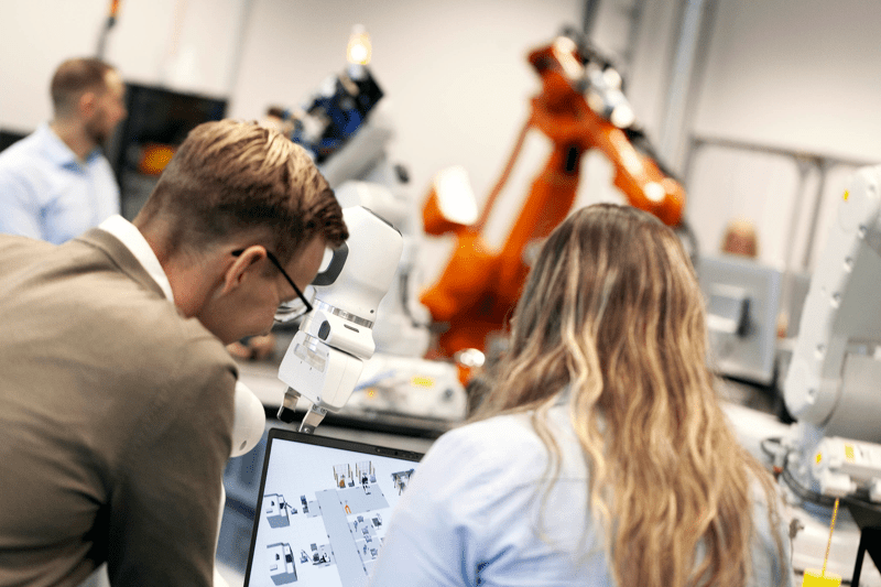 Two engineers working together at a workstation in an industrial lab with robotic arms in the background