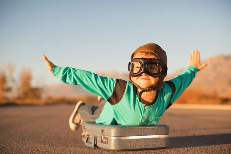 A young boy lies on top of a suitcase with his arms outstretch as if flying. He wears a flight cap and goggles. 