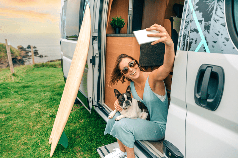 Woman and dog taking a selfie, sat in a camper van overlooking a beach. 