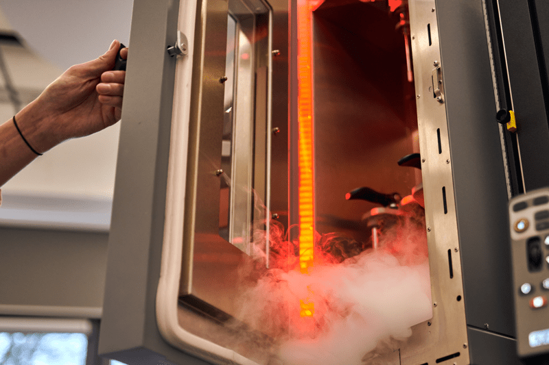 A hand opens the door of a laboratory testing chamber filled with red interior lighting and emitting vapor.