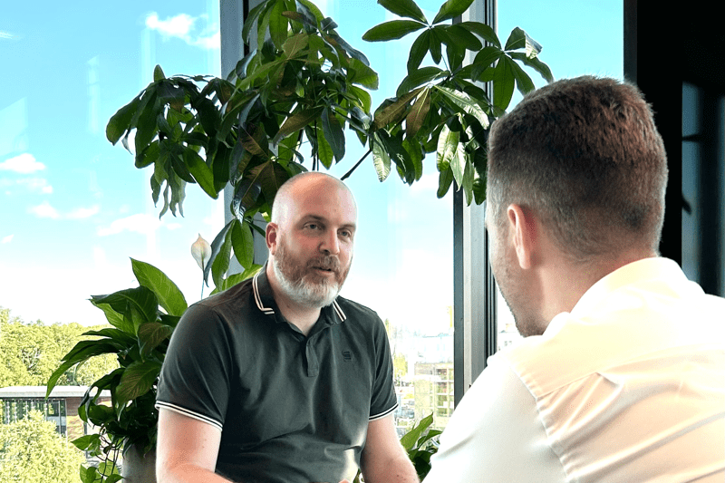 Man i grå pikétröja ler och samtalar med en person i vit skjorta som har ryggen vänd mot kameran. I bakgrunden syns grönväxter framför fönster.