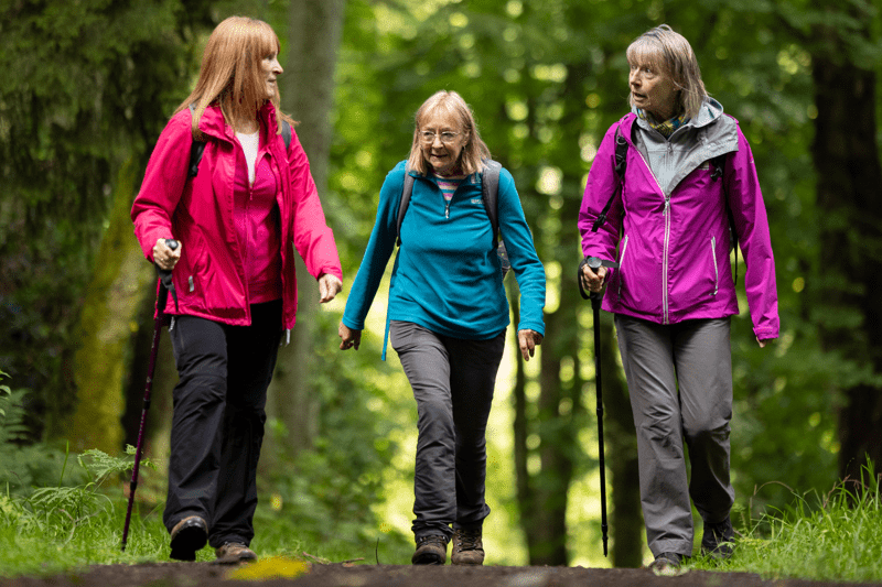 Three people wearing colourful outdoor jackets walk along a woodland path surrounded by green trees. Text overlay reads “Our Benefits”.