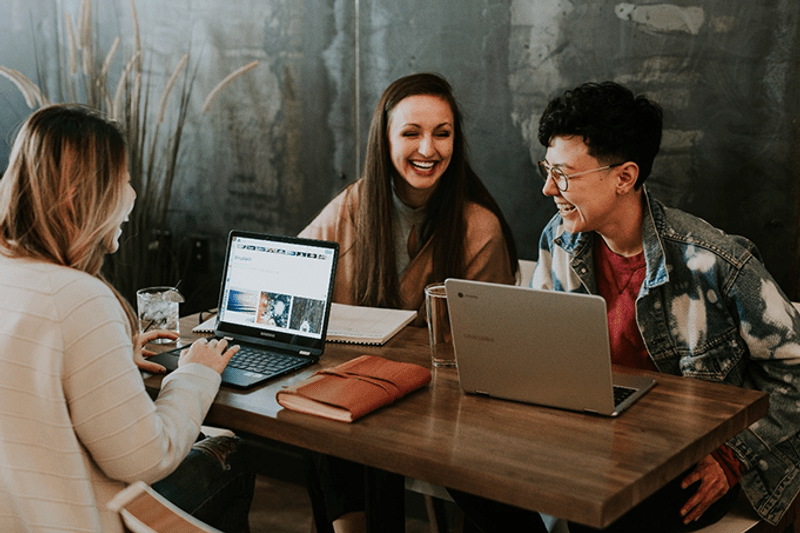 Three young adults laugh and talk while working on laptops at a wooden table in a cafe, with a leather notebook and drinks nearby.