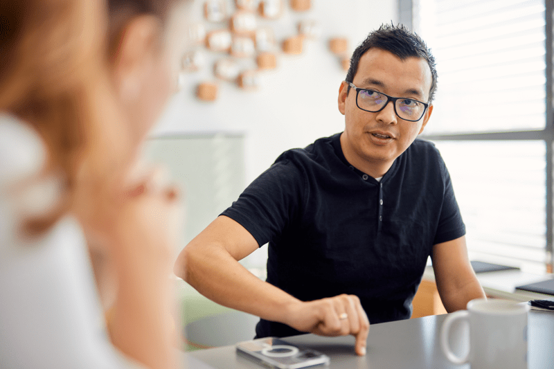 Man in glasses engaged in conversation with a colleague across a desk in a modern office