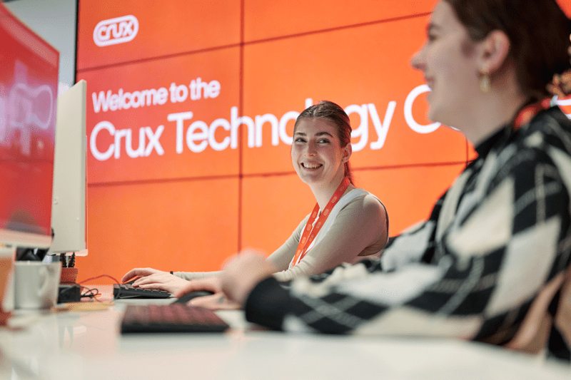 Two women smiling in the foreground, with orange screens behind them displaying ‘Welcome to the Crux Technology Centre’ in white text.
