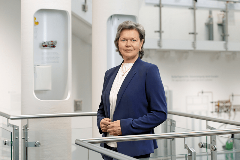 Elena Skvortsova wearing a navy blazer and white shirt standing indoor the industrial museum near a glass railing with white cabinets in the background