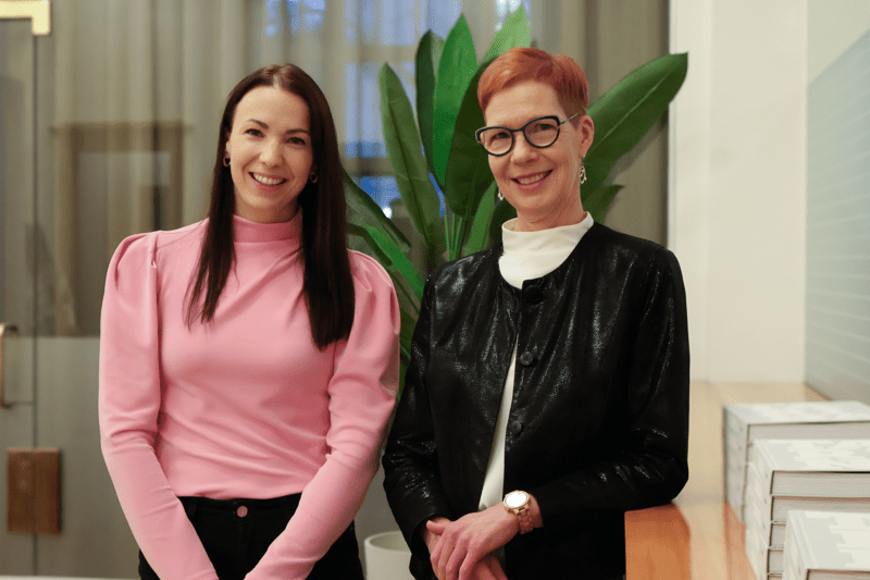 Kaksi työntekijää seisoo rinnakkain toimistollaan katsoen kohti hymyillen. 