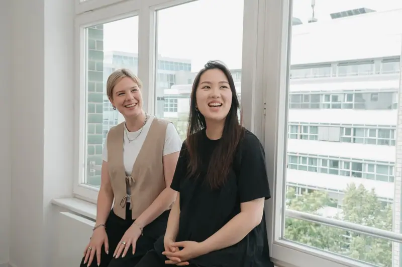 Two colleagues sitting together by a window, smiling in a bright office environment.