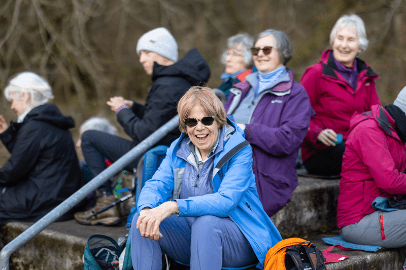 A group of six people sitting on stone steps in a wooded area, with one woman in a blue jacket smiling in the foreground. Text overlay reads "member and supporter experience"