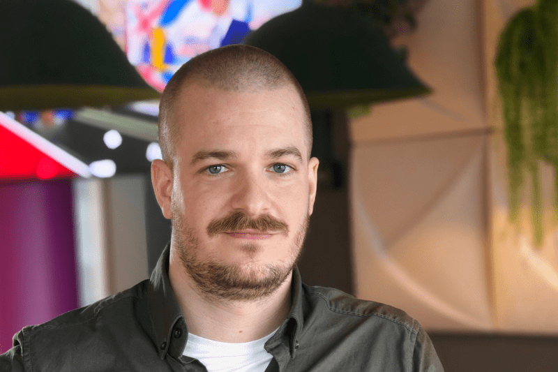 Leende man med rakat huvud och kort skägg, iförd grå skjorta över vit t-shirt, står i ett färgstarkt kontorsutrymme med moderna möbler och hängande lampor i bakgrunden