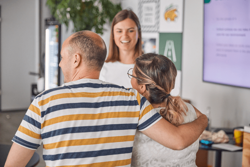 Three people smiling and greeting each other warmly in an office, with one person placing an arm around another