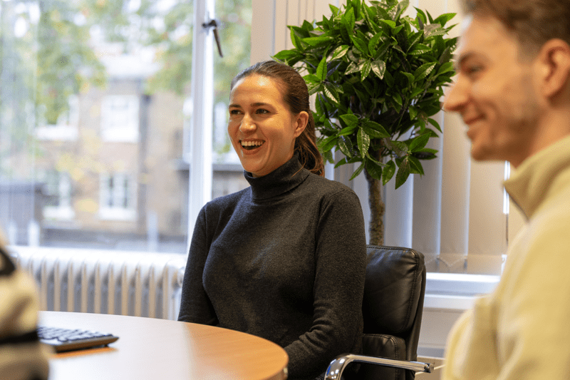 Woman laughing during a meeting, seated at a round table beside a window with a potted plant behind her.