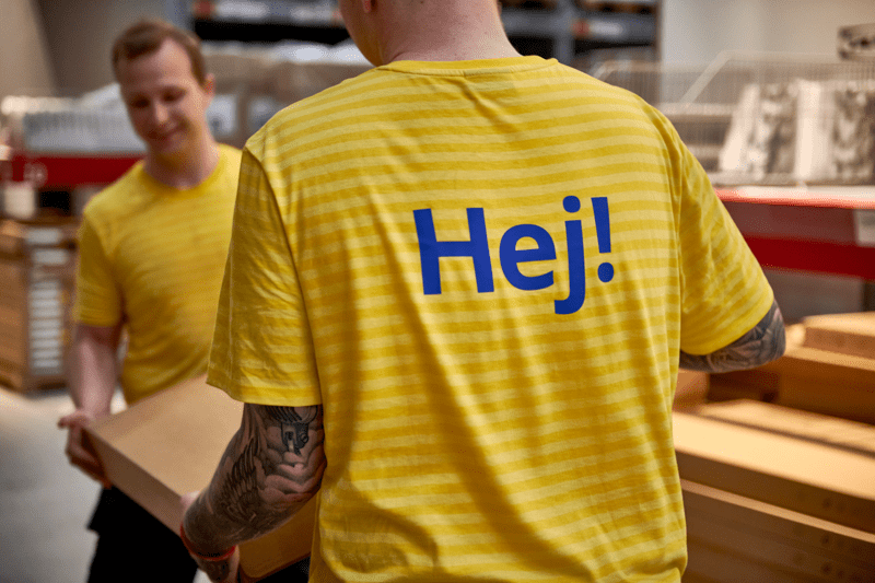Du IKEA darbuotojai su dryžuotomis geltonos spalvos uniformomis ir užrašu „Hej!“ ant vieno iš jų nugaros kartu neša kartoninę dėžę prekių užsakymo ir atsiėmimo vietoje.