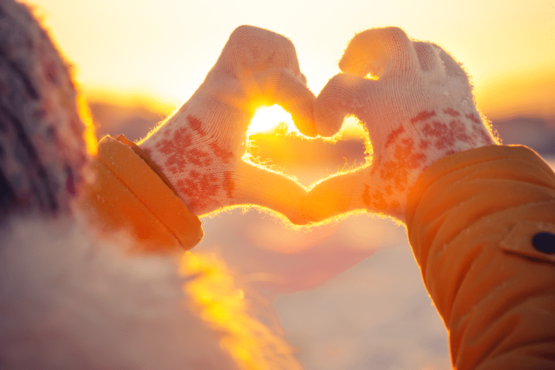 A child holding their gloved hands in a heart shape, against a sunset backdrop