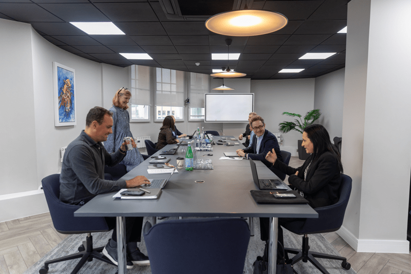 Colleagues seated around a long meeting table with laptops, talking and preparing for a meeting.