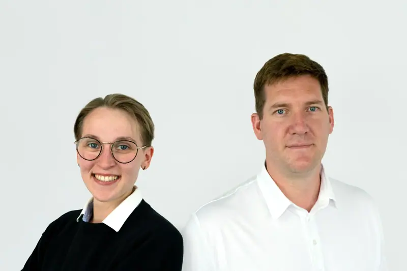 Two colleagues standing side by side against a light background, smiling at the camera.