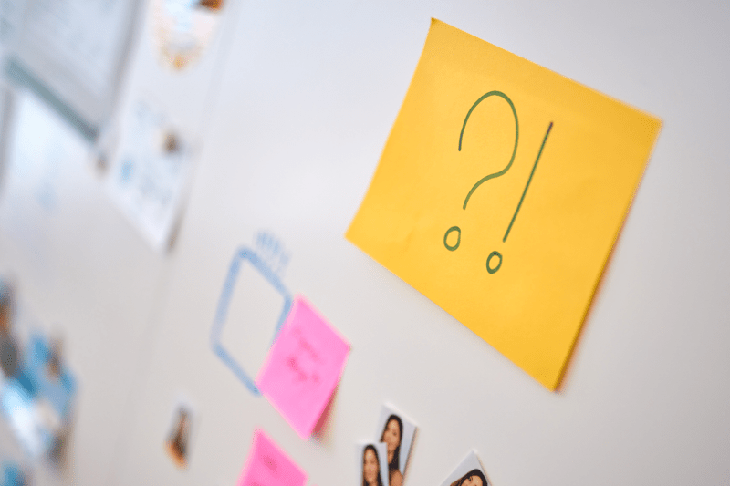 Yellow sticky note with a question and exclamation mark on a whiteboard, surrounded by other colorful notes and images
