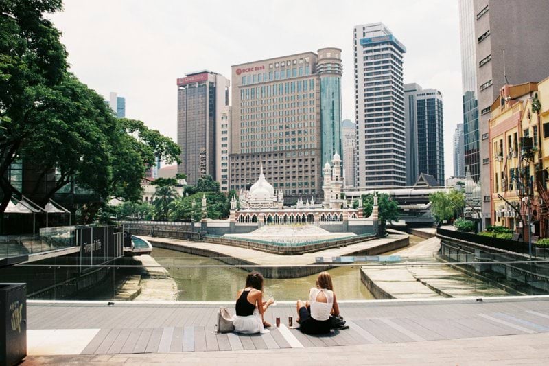 two women sitting on concrete pavement facing on mountain during daytime