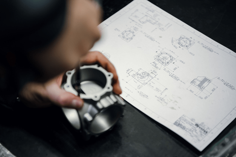 Person holding a mechanical part over an engineering drawing