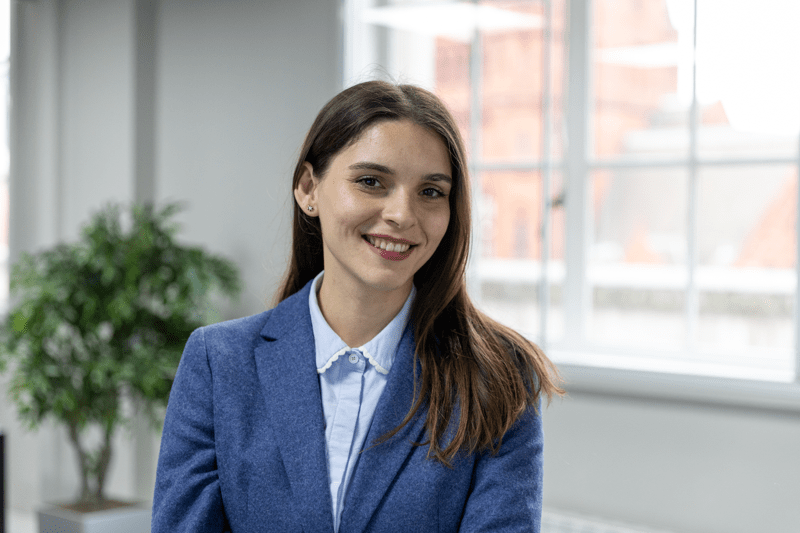 Woman in a blue blazer smiling in a bright office with large windows behind her