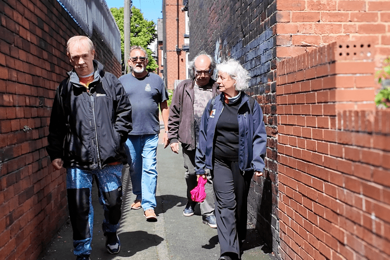 Several people walk together along a brick-lined passageway in an urban setting. Text reads: “Delivery - England”.