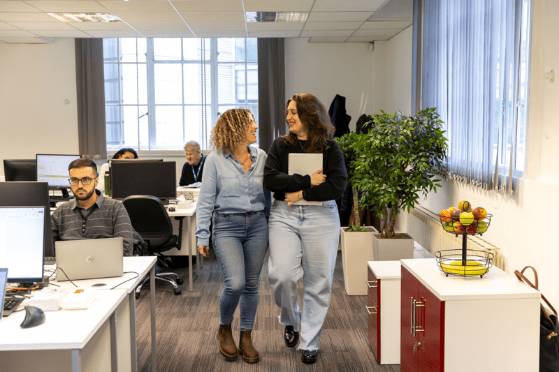 Two colleagues walking through an open-plan office, smiling and talking, with other staff working at desks behind them.
