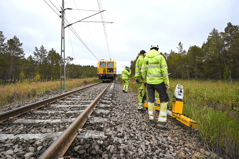 Tre män står vid en järnväg under ett rälsarbete. I bakgrunden ser man ett spårriktar-tåg.