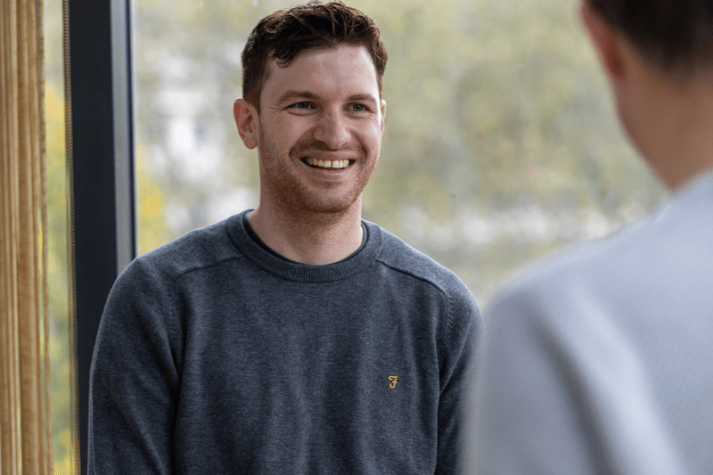 Man in a grey jumper smiling while speaking with someone off-camera near a window.