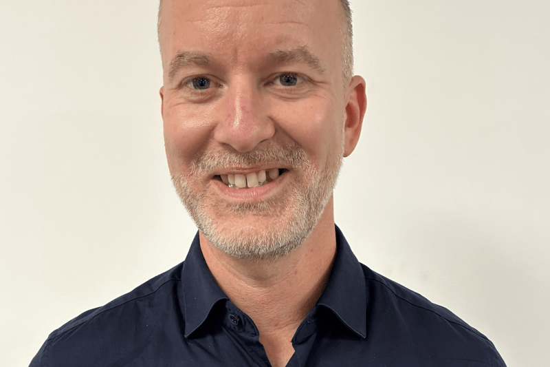 ersonen står mot en ljus vägg med en gul kudde synlig bakom axeln. Enkla, neutrala färger och fokus på överkroppen.