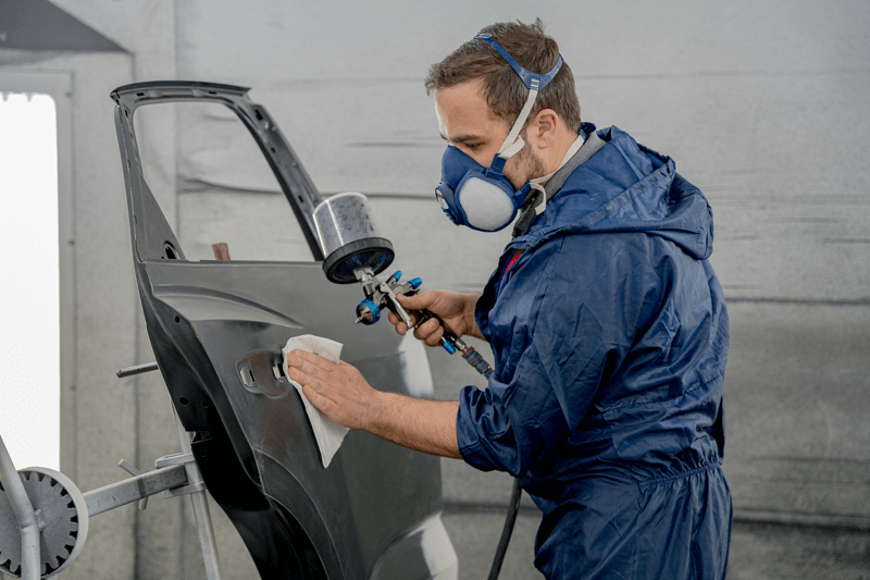 Carrossier portant une blouse et un masque de protection, effectuant des gestes minutieux pour travailler avec précision sur la voiture.