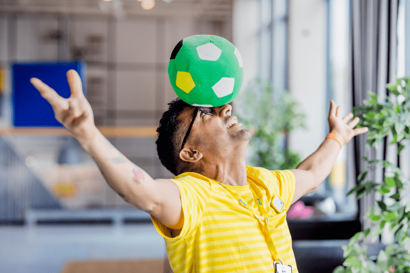 IKEA darbinieks dzeltenā uniformā, pacēlis rokas rotaļīgā pozā, uz galvas balansē mīkstu, zaļu futbola bumbu.
