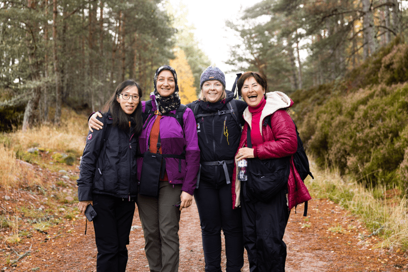 Four people wearing outdoor jackets and backpacks stand together on a woodland path, with trees and heather-covered ground surrounding them. Text overlay reads “Delivery - Scotland”.