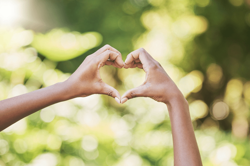 	Två händer formar ett hjärta mot en suddig, grön bakgrund utomhus.