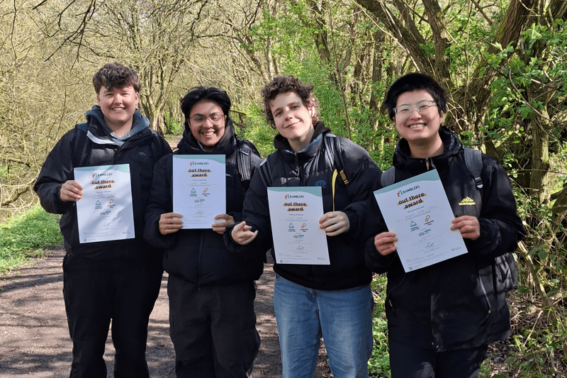 Four people stand on a woodland path holding certificates, wearing outdoor clothing and backpacks. Trees and greenery surround the group. Text overlay reads “Programmes”.