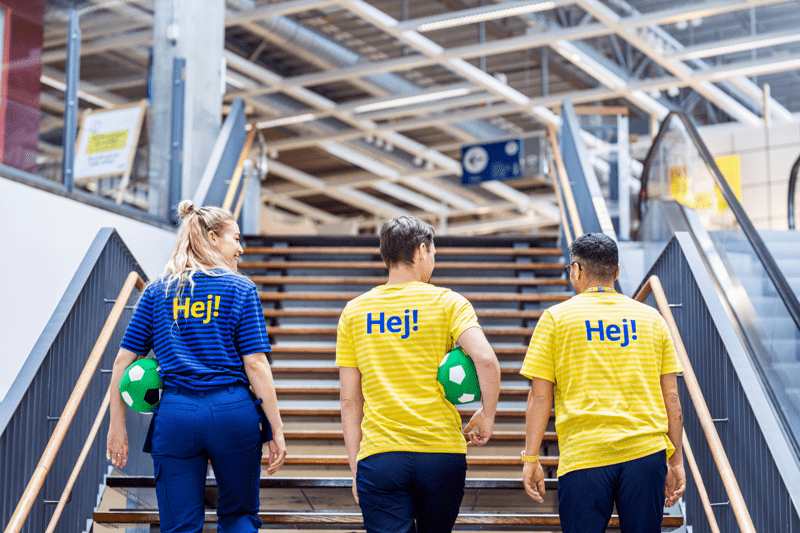 Trīs IKEA darbinieki dzeltenos un zilos T-kreklos ar uzrakstu „Hej!” uz muguras kāpj pa veikala kāpnēm.