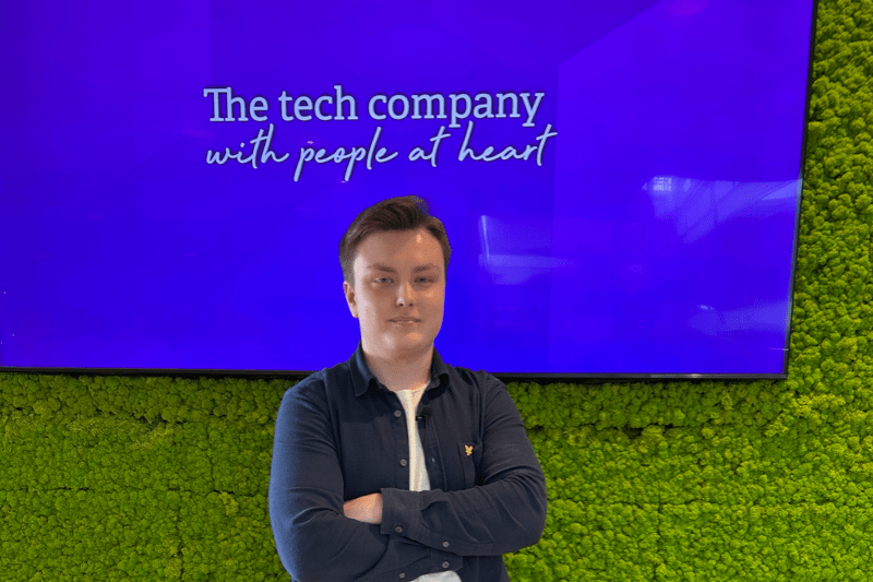 Ung man med mörkblå skjorta och vit t-shirt står framför en grön vägg med stor lila skärm. På skärmen står texten: 'The tech company with people at heart'.