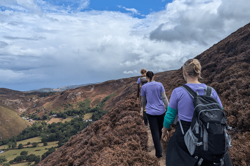 Three people wearing backpacks walk along a narrow hillside path overlooking a valley with rolling hills and rocky slopes. Text overlay reads “Delivery - Wales”.