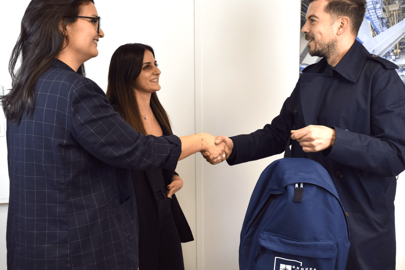 Two women welcome a man into an office. One of them shakes his hand while the other stands beside her. The man is wearing a coat and carrying a blue backpack with the ADF logo.
