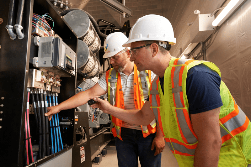 An inspection employee wearing an orange vest and white hard-hat alongside a contractor employee wearing wearing a similar vest and hat look at electrical equipment in a brightly-lit area under construction.