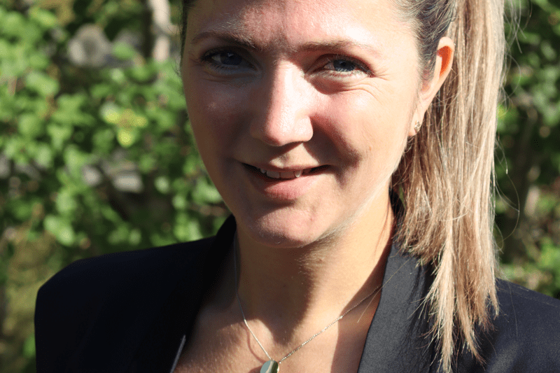 Närbild på medarbetare i mörk kavaj och uppsatt hår, natur i bakgrunden.. Tittar rakt in i kameran