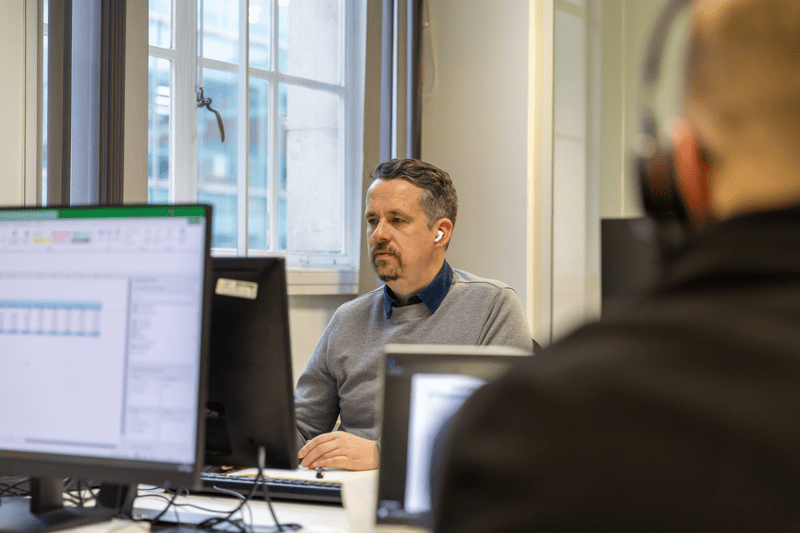 Man wearing wireless earbuds working at a desktop computer in an office near a window.