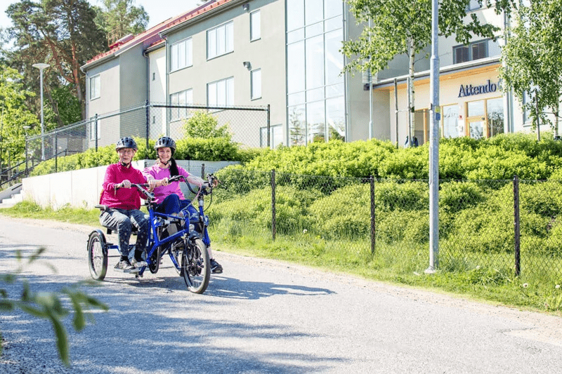 En omsorgsmedarbetare och en kund cyklar på en slags tandemcykel, där de båda sitter bredvid varandra. 