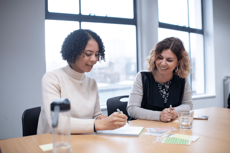 Two Anthony Nolan employees sat side-by-side in a meeting room, one is writing notes