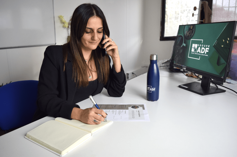 Een medewerkster van Groupe ADF voert een telefoongesprek aan haar bureau terwijl zij aantekeningen maakt in een notitieboek. Voor haar liggen een geprint cv en een scherm waarop het logo van Groupe ADF wordt weergegeven.