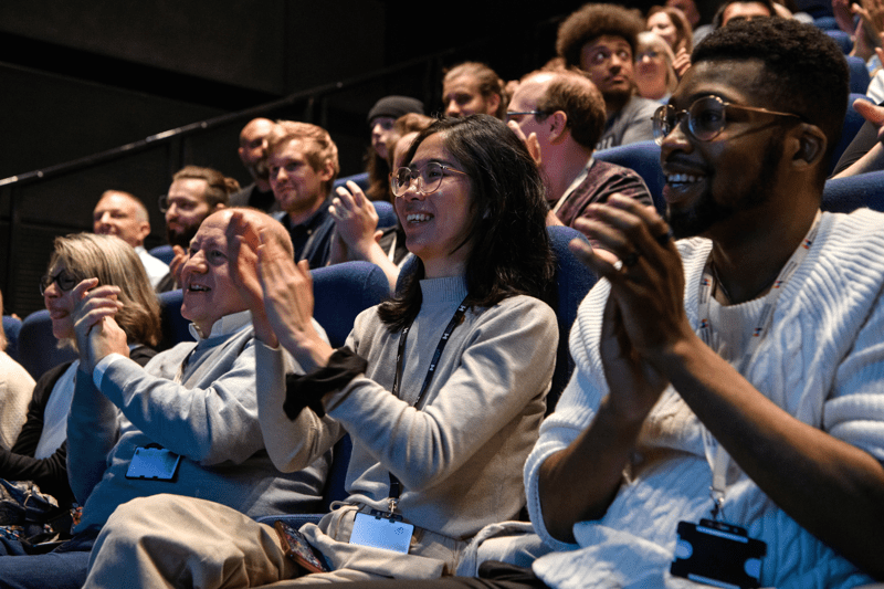 Staff members sitting in an auditorium clapping at something onstage.