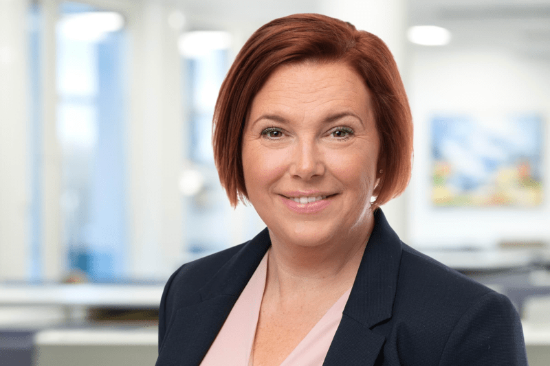 Kvinna i kavaj ler mot kameran i ljus kontorsmiljö med suddig bakgrund.