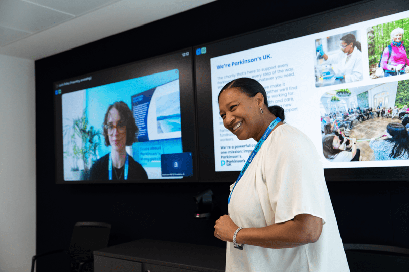 A colleague presenting to employees in a meeting room, with one employee joining virtually on screen.