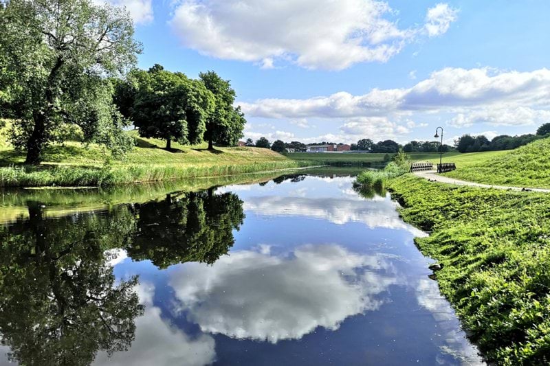 green grass field and trees beside river under blue sky and white clouds during daytime