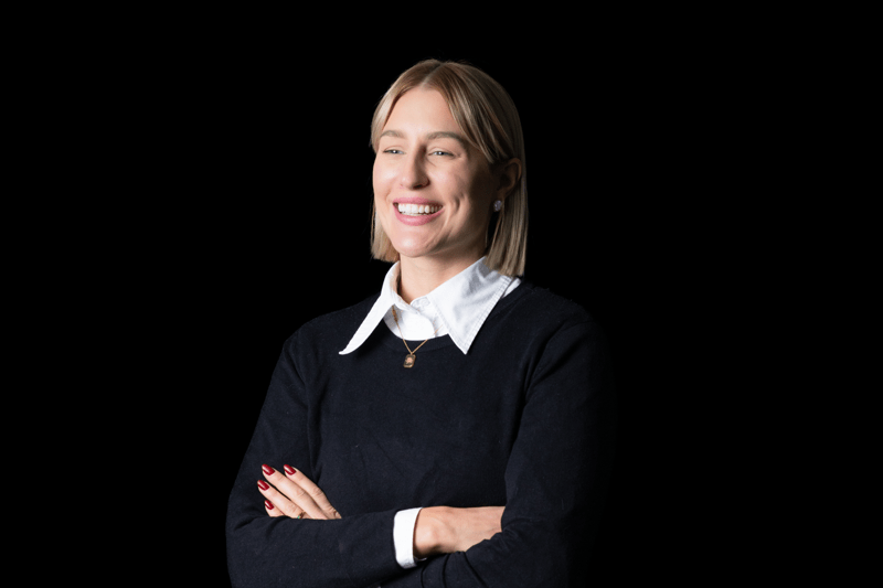 A smiling woman with short blonde hair stands against a black background, wearing a black sweater over a white collared shirt, with her arms crossed.